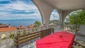 Ein Balkon mit rotem Tischläufer, einer hölzernen Bank und Blick auf das Meer und die Bebauung.