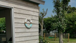 Une maison de plage en bois, avec une enseigne indiquant « Maison de plage » et deux bannières décoratives sur la terrasse.