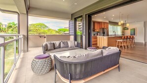 Ein moderner Wohnbereich mit einer Couch, einem Hocker und einem Esstisch. Durch eine offene Tür ist eine Küche erkennbar, und ein Balkon bietet Blick auf eine Grünfläche.