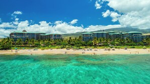 Ein Strandhotel mit mehreren Gebäuden, klarem türkisblauem Wasser und einem Sandstrand mit Sonnenschirmen und Personen.