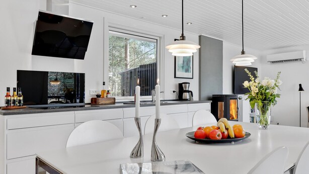 Una cocina moderna con muebles blancos, un ventanal grande, un televisor de pantalla plana y un frutero en la isla.