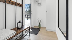 Un interior moderno con un banco de madera, una mesa de cristal, una estantería de metal, un tapete negro y una puerta de madera que da al exterior.