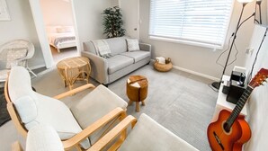 Una sala de estar con un sofá gris, una silla de madera, una mesita redonda, una guitarra y una ventana con persianas.