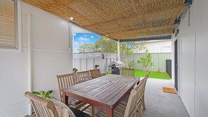 Un patio cubierto con una mesa y sillas de madera, una parrilla y vistas a un jardín trasero con cerco y follaje.