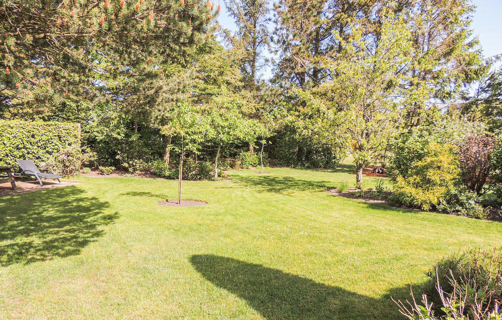 Um jardim bem cuidado com diversas árvores e arbustos, gramado e um banco de madeira.
