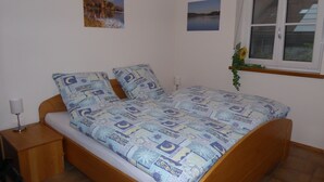 A bedroom with a bed, a nightstand, a lamp, a window with a plant, and two wall-mounted pictures.
