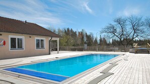 En rektangulär swimmingpool omgiven av ett trädäck, ett takförsett sittområde och en byggnad med ett tegeltak i bakgrunden.