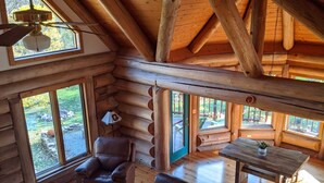 Ein großzügiges Blockhaus-Interieur mit hoher Decke, Holzbalken, großen Fenstern, einem Ledersofa und einem Esstisch.