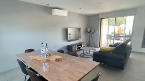 Uma sala de estar moderna com área de jantar, destacando uma mesa de madeira, cadeiras pretas, um sofá cinza e uma TV fixada na parede.
