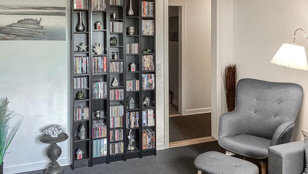 A room with a bookshelf filled with books, a grey armchair, a small table with a vase, and a lamp. - Gorgeous apartment in Rødekro with WiFi (Rødekro)