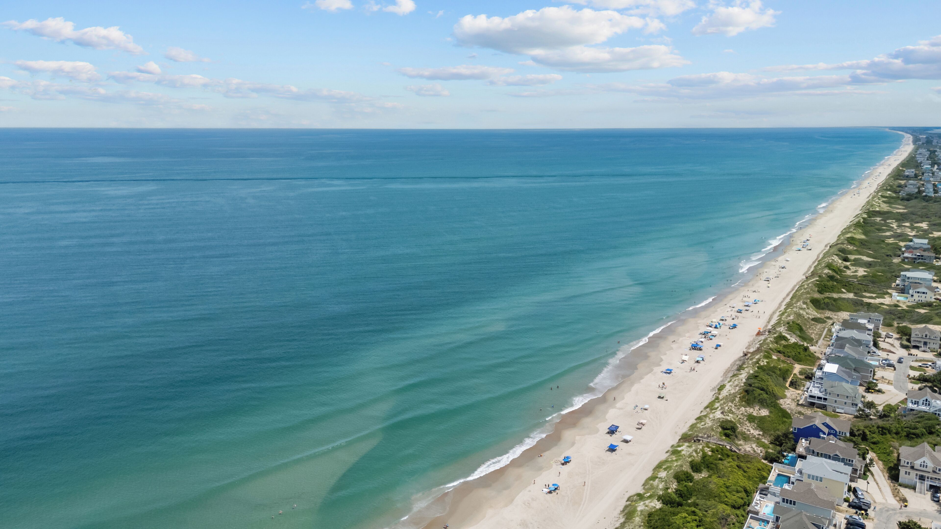 An aerial view of a coastal area with a beach, houses, and a clear sky.