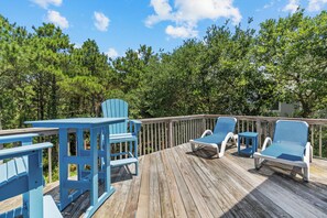 A wooden deck with blue Adirondack chairs and a table, surrounded by trees.