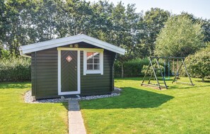 Situated in a serene yard, this charming shed features a gabled roof and a front door, surrounded by lush greenery and a wooden swing set.