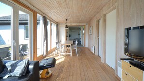 Una habitación amplia con suelo de madera, un televisor grande a la derecha, una zona de comedor en el centro y ventanas grandes a la izquierda.