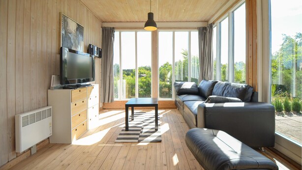 Un salón moderno con un televisor grande, suelo de madera, un sofá y vistas a la vegetación a través de amplios ventanales.