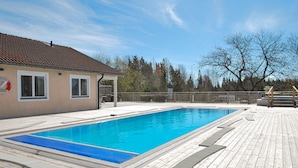 A rectangular swimming pool with a wooden deck surrounding it, a covered seating area, and a building with a tiled roof in the background. - 1 bedroom beautiful home in Kosta (Kosta)