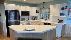 A modern kitchen with white cabinetry, a stainless steel refrigerator, a microwave, and a gas stove. There is a window with a view of trees, and a laptop on the countertop.