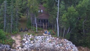 A cabin surrounded by dense forest, a stone wall, and a small dock extending into a body of water.