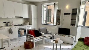 En kompakt opholdsstue med tekøkken, spiseplads, siddeområde med stol og skammel, et fjernsyn og et vindue med skodder.