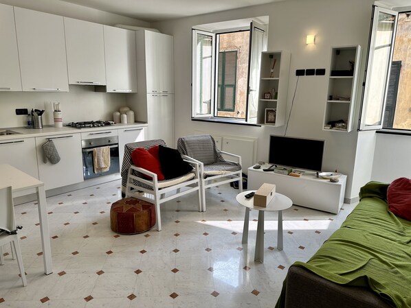 Un espace de vie compact comprenant un coin cuisine, un coin repas, un coin salon avec un fauteuil et un pouf, un téléviseur et une fenêtre avec des volets.