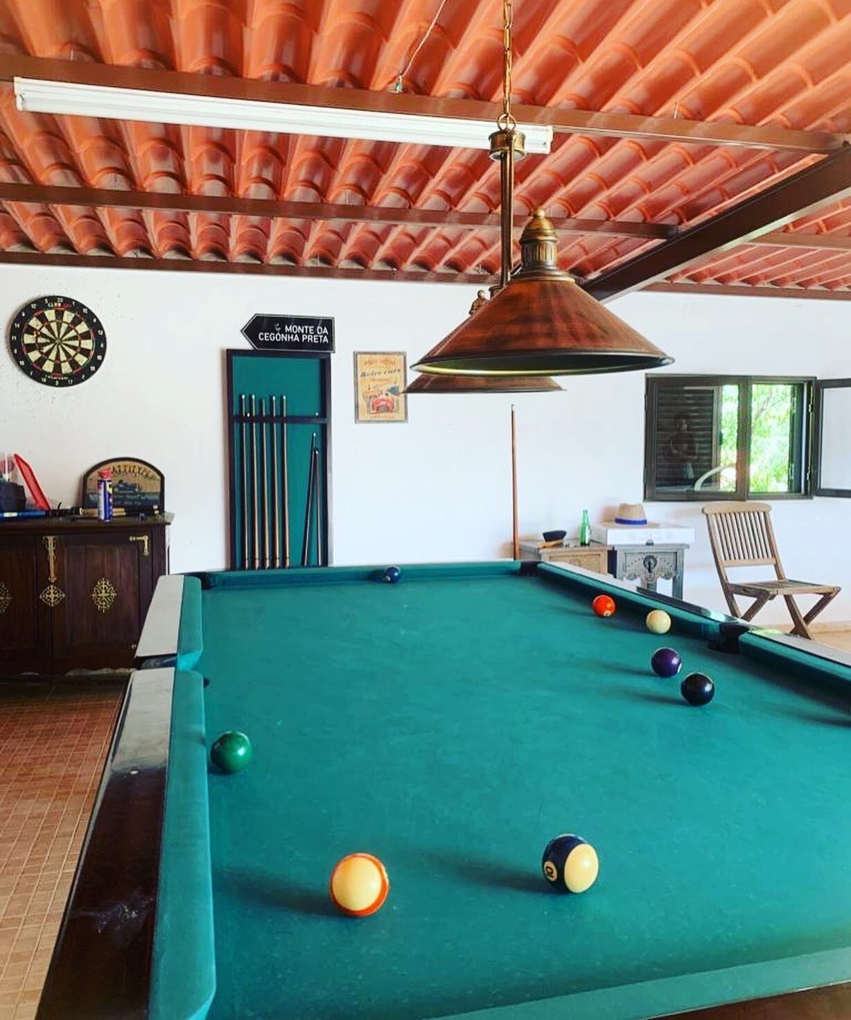 A pool table with balls, a dartboard, and a billiards cue rack in a room with a tiled ceiling and a window.