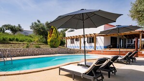 A pool area with lounge chairs and umbrellas, a stone wall, and a building with a blue and white facade.