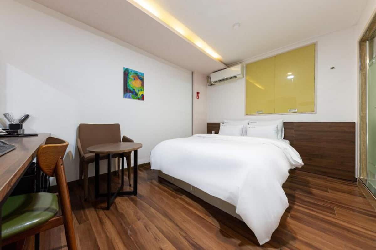 A hotel room with a wooden floor, a bed with white bedding, a desk with a chair, and a large window.
