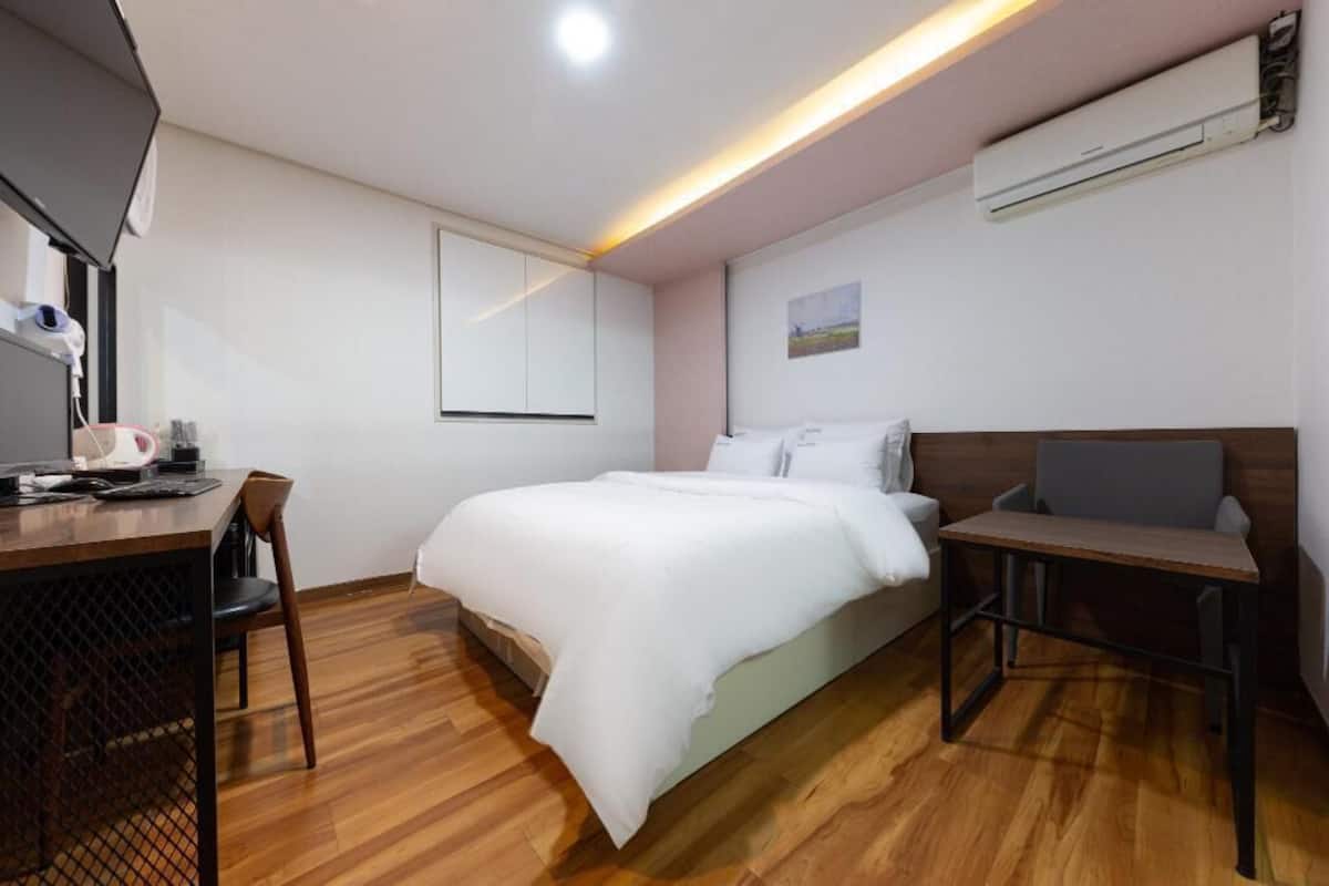 A modern hotel room with a wooden floor, a bed with white bedding, a desk with a chair, and an air conditioning unit on the wall.