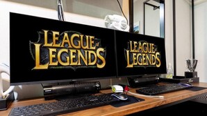 Two monitors displaying 'League of Legends' on a wooden desk with a keyboard, mouse, and a small trophy. - NJ Hotel (Pyeongtaek)