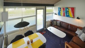 Ein modernes Wohnzimmer mit einer braunen Ledersofa, einem weißen Couchtisch und einem gelb-weißen Teppich. Zwei Fenster mit Jalousien, ein Heizkörper und eine Deckenleuchte sind vorhanden.