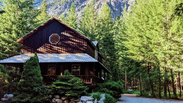 A two-story log cabin with a metal roof, surrounded by dense forest and mountains in the background. - Cozy Mazama Studio on 5 Acres-Trail Access & Views (Mazama)