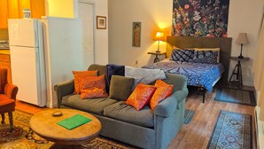 A compact living space with a sofa, a small round table, a bed with patterned bedding, a tapestry on the wall, and a kitchen area with a refrigerator. - Cozy Mazama Studio on 5 Acres-Trail Access & Views (Mazama)