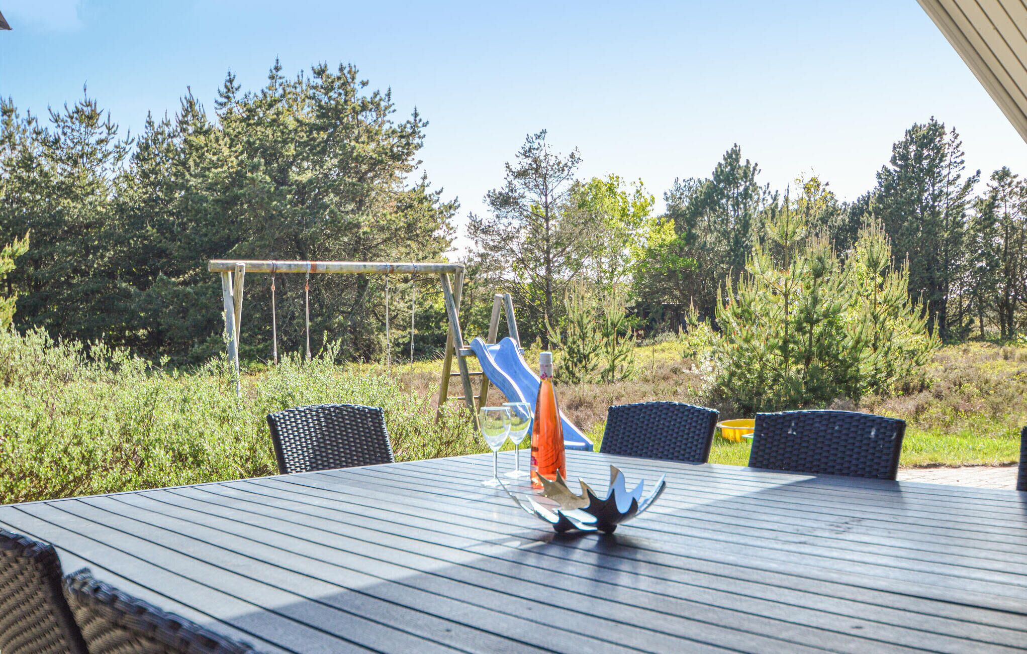 Uma área de refeições ao ar livre com mesa e cadeiras, uma peça de metal decorativa e um escorrega azul ao fundo.