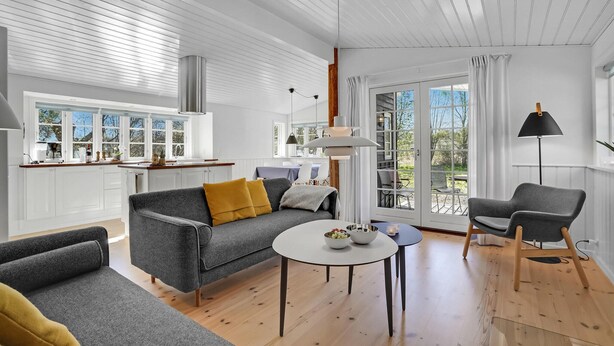 A modern living room with a grey sofa, a round coffee table, and a grey armchair. There is a kitchen area with white cabinets and a window overlooking the outdoors. - 3 bedroom gorgeous home in Glesborg (Glesborg)