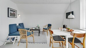 A modern, well-lit room with a white ceiling, a dining table with chairs, a sofa, and a television mounted on the wall.