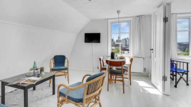 A modern, well-lit room with white walls and a sloped ceiling. It features a dining table with chairs, a television mounted on the wall, and large windows offering a view of the cityscape.