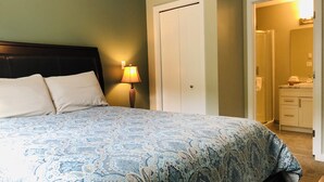 Een slaapkamer met een bed, een nachtkastje met een lamp, een deur en een zichtbare badkamer door een geopende deur.