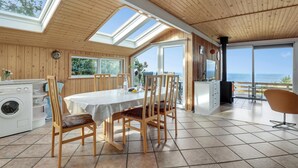 A cozy kitchen with wooden walls, a dining area with a table and chairs, a washing machine, and a large window overlooking the sea. - Stunning home in Hasle with WiFi (Hasle)