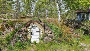 Un arc en pierre rustique mène à une porte en bois, le tout étant entouré d’un feuillage dense et d’un petit bâtiment doté d’un toit en tuiles.