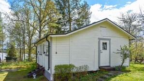 Une petite cabane blanche d ’un seul étage, avec une porte d’entrée et des fenêtres, entourée d’arbres et d’herbe.