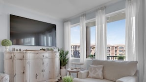 A living room with a white sofa, a white cabinet, a flat-screen TV, and a balcony with a view of buildings. - Any day at the beach is a good day! 🏖️ Book your vacay today!  (Miramar Beach)