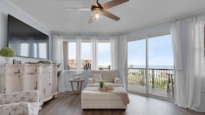 A spacious living room with a large window, a ceiling fan, and a view of the sea. - Any day at the beach is a good day! 🏖️ Book your vacay today!  (Miramar Beach)