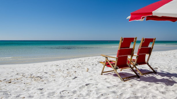 A beach scene with two red beach chairs and a red and white beach umbrella. - Any day at the beach is a good day! 🏖️ Book your vacay today!  (Miramar Beach)