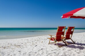 A beach scene with two red beach chairs and a red and white beach umbrella. - Any day at the beach is a good day! 🏖️ Book your vacay today! (Miramar Beach)