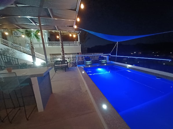 A rooftop pool area with a blue-lit pool, outdoor seating, and a covered terrace.