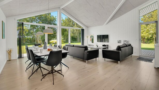 A spacious room with a dining table, black chairs, a sofa, and a TV. Large windows provide a view of the outdoors.