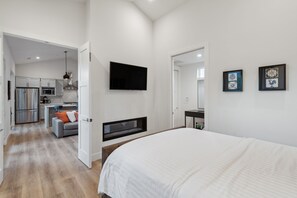 Ein modernes Schlafzimmer mit einem großen Bett, einem flachbildfernseher an der Wand und einer kleinen Küchenzeile im Hintergrund.