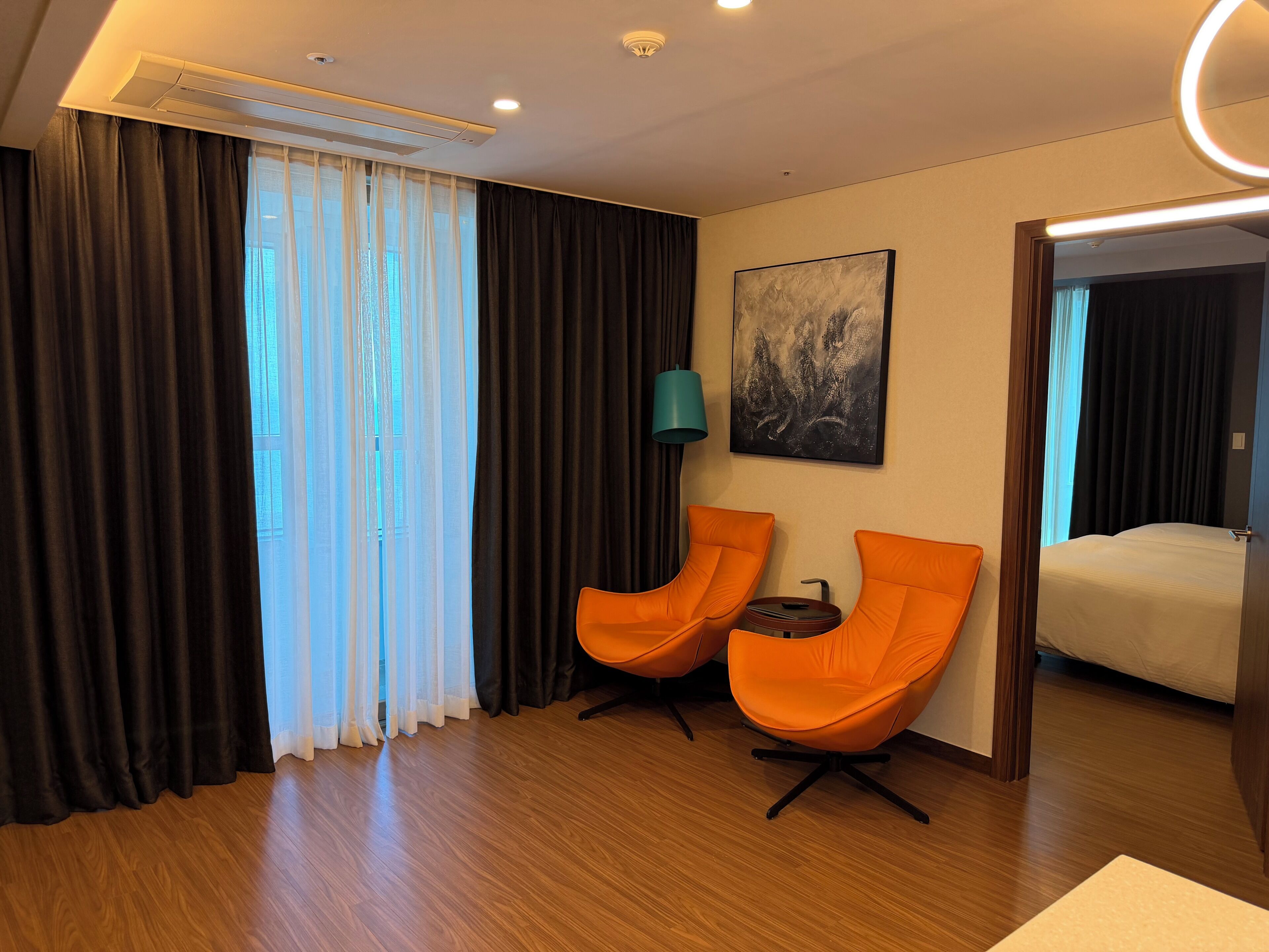 Ein Hotelzimmer mit zwei orangenen Sesseln, einem Bett, einem Bild an der Wand und einem Fenster mit Vorhängen.