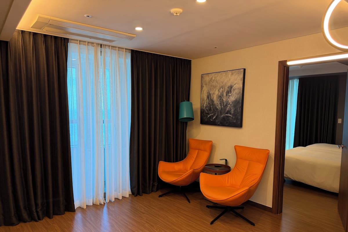 Ein Hotelzimmer mit zwei orangenen Sesseln, einem Bett, einem Bild an der Wand und einem Fenster mit Vorhängen.
