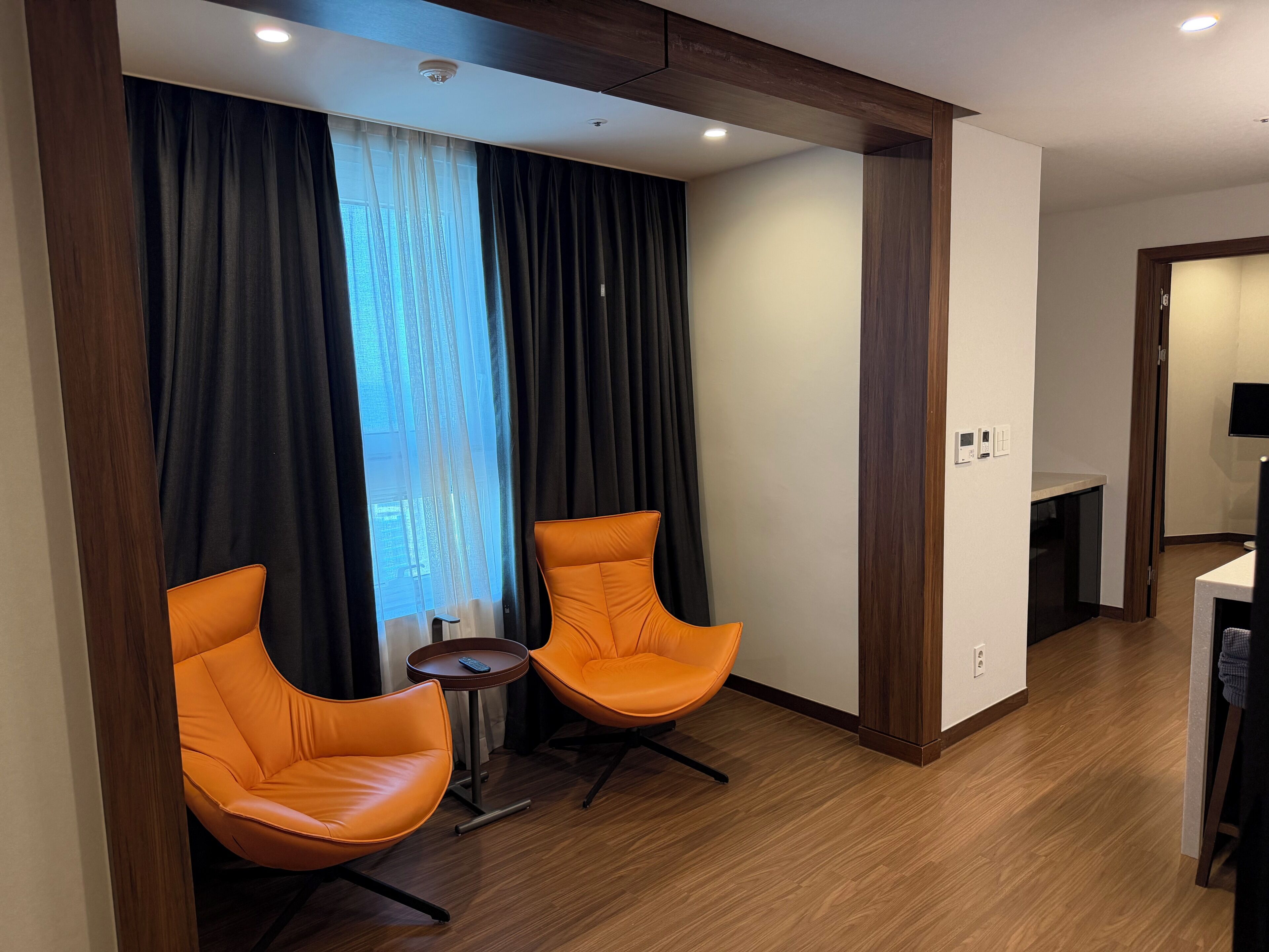 Ein modernes Hotelzimmer mit zwei orangenen Sesseln, einem kleinen Tisch, einem Fenster mit Vorhängen und einem Holzboden.